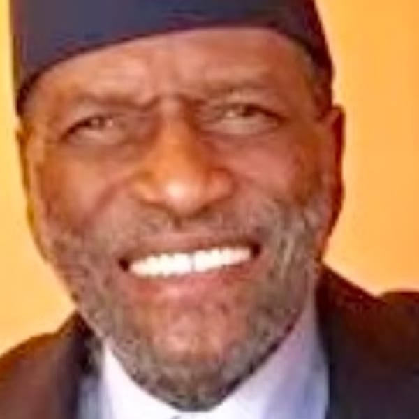 Close-up of a smiling man with a beard and a dark cap, against an orange background.