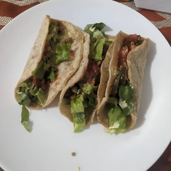 Three vegan tacos filled with lettuce, tomatoes, and a creamy sauce on a white plate.
